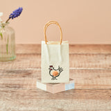 A small cream-colored paper gift bag with twisted kraft paper handles, featuring a cute embroidered chicken design on the front. The bag is displayed on a pastel-colored geometric block on a rustic wooden table, with a blurred glass vase in the background.