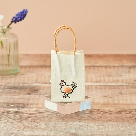A small cream-colored paper gift bag with twisted kraft paper handles, featuring a cute embroidered chicken design on the front. The bag is displayed on a pastel-colored geometric block on a rustic wooden table, with a blurred glass vase in the background.